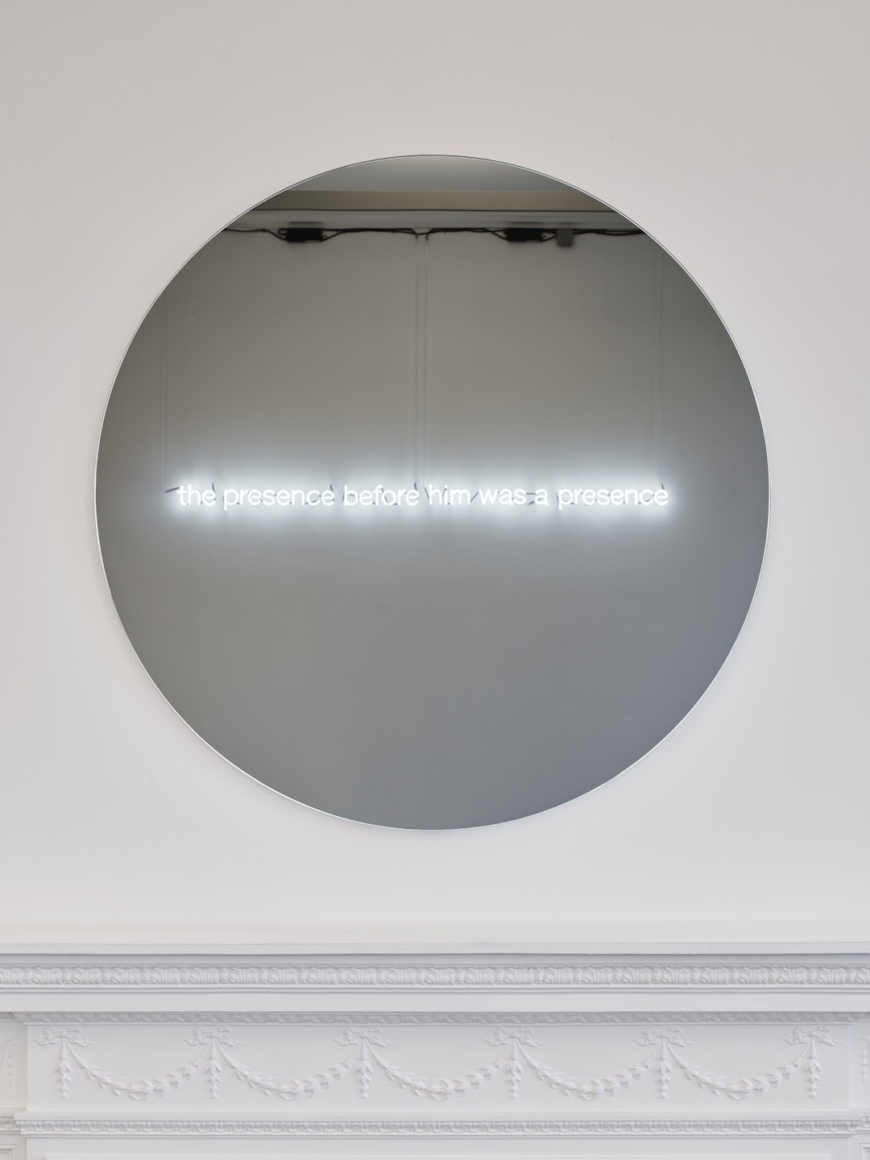 Walker &amp; Walker, The presence before him was a presence (Henry James) is a neon text written in reverse, continuing Walker and Walker’s series of works fabricated in neon. These works are often hung facing windows or only seen clearly as a reflection. In this case, viewing the neon sign in the black mirror opposite it reveals the words ‘the presence before him was a presence’. This phrase is taken from The Jolly Corner, a short story by the American writer Henry James in which the protagonist is confronted with a ghostly alter ego. This ghost represents the different paths he could have taken in his life, and consequently, the different people he might have become. In Walker and Walkers work, their concern is to suggest a presence that accompanies the viewer throughout the exhibition, oscillating between the material and immaterial. To view the work one turns away from its physical manifestation, to a reflection in a mirror, augmenting the sentiment of the work.