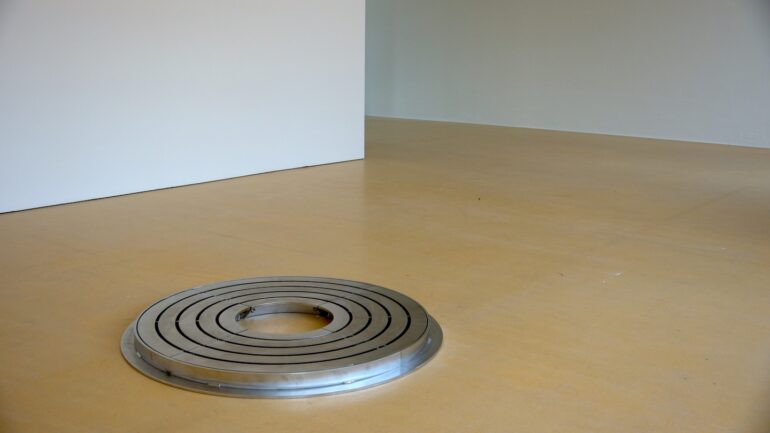 Walker &amp; Walker, IN WAITING (OAK TREE), 2015
Stainless steel, 96cm diameter x 5 cm 
Accompanying these works, a stainless-steel circular sculpture adhering to the language of minimalism is placed on the floor. Almost an ubiquitous object, it is a tree holder or a protective guards usually seen on some city streets. Picking up on the motif of zero which addresses the notion of absence, loss, or the void, this object is in essence is designed in time to be obsolete; as the tree outgrows its designated circle, each defined space is removed in turn. This work identifies another ongoing theme within Walker and Walker’s work; the tension between presence and absence. ‘In waiting (Oak tree)’ is an object filled with the potentiality of the physicality of a tree but is also filled with a certain redundancy, as it sits seemingly in a state of exile.